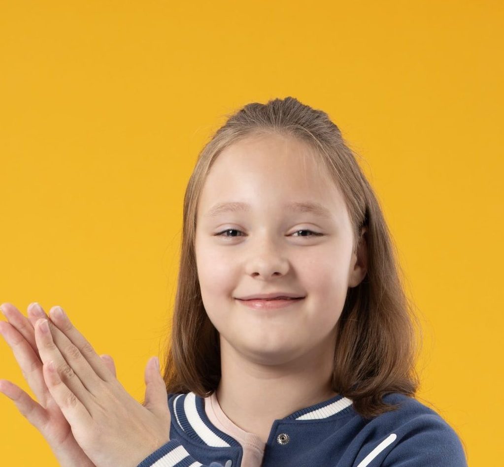 Zdjęcie przedstawia portret Julii Miłek, uśmiecha się i klaszcze w dłonie. Ma włosy do ramion. Całość na żółtym tle.