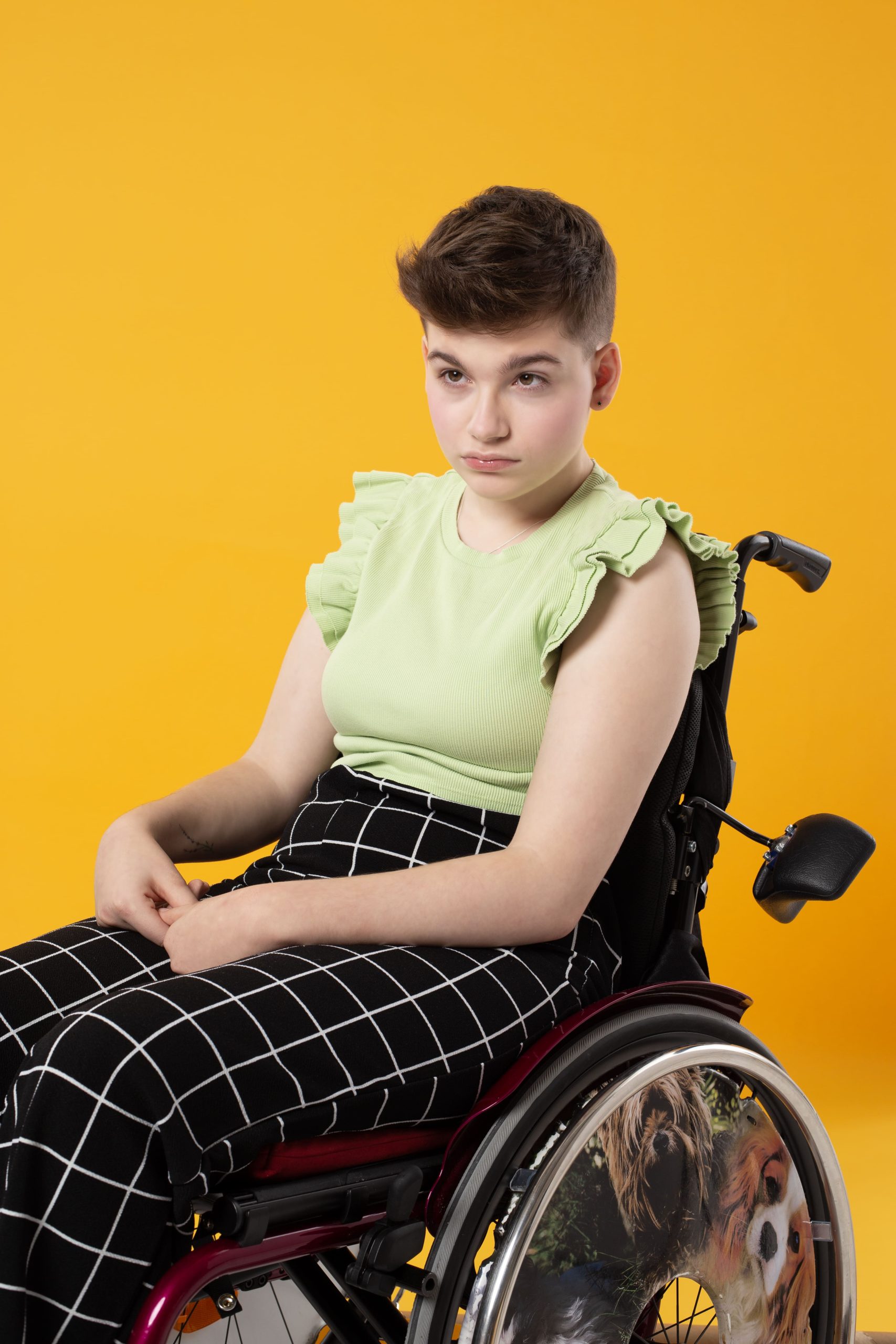 Zdjęcie przedstawia Gabrysię Miszta o krótko ściętych włosach ułożonych do góry, w spodniach i bluzce siedzącą na wózku ręcznym. Całość na żółtym tle.