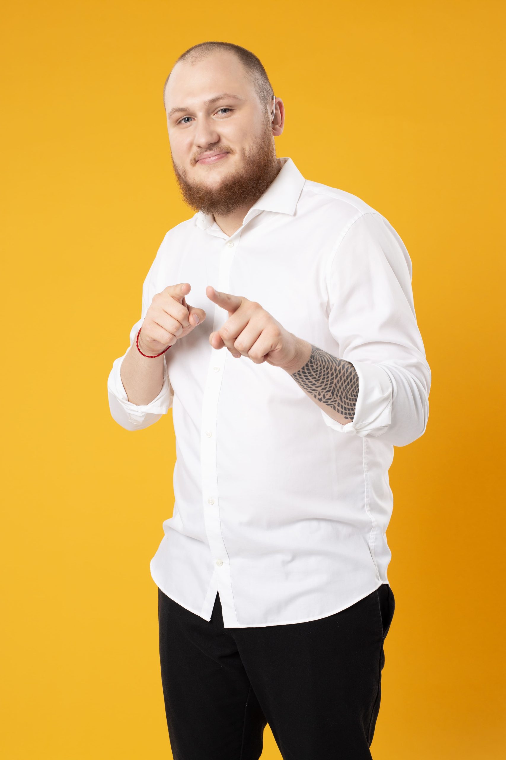 Zdjęcie przedstawia Jakuba Stanisławczyka w białej koszuli porozumiewawczo wskazuje palcami wskazującymi. Całość na żółtym tle.