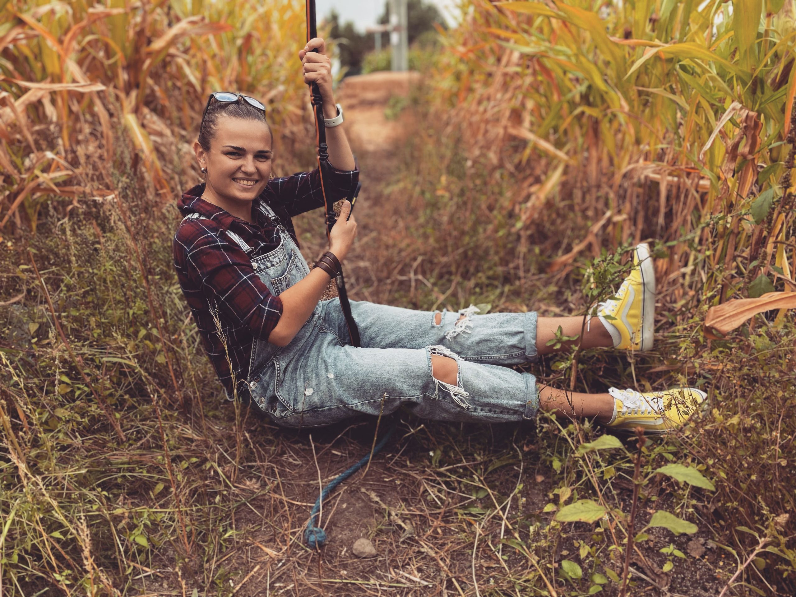 Zdjęcie przedstawia uśmiechniętą Olesię Vasichkina w polu kukurydzy na huśtawce ze sznura ubrana jest w brązową koszulę i ogrodniczki z dziurami