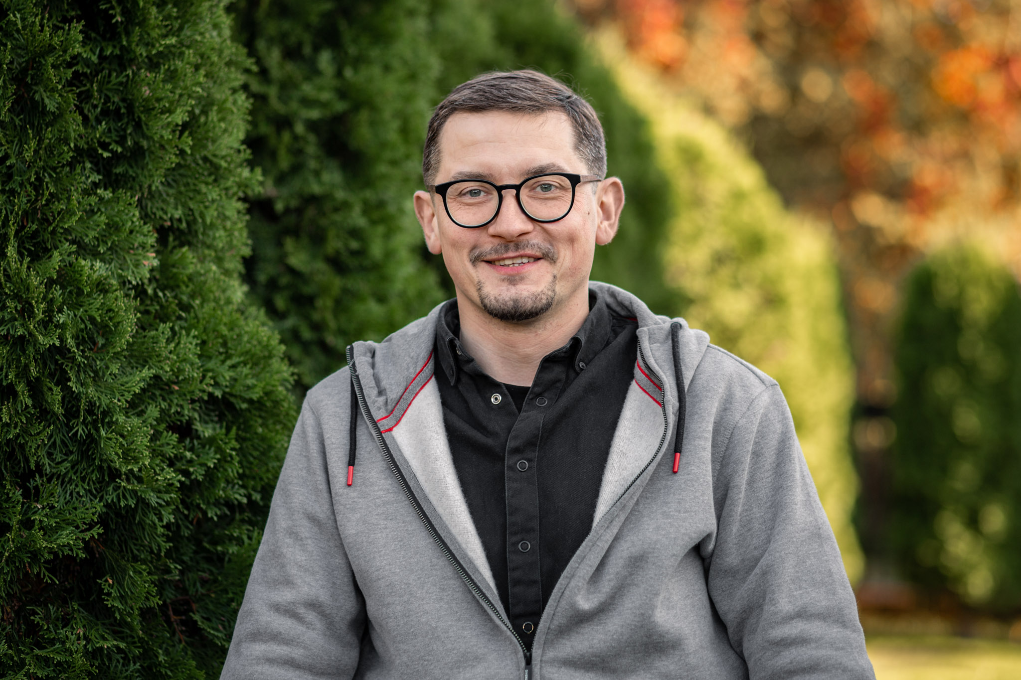 Portret Tomasza Grabowskiego w szarej bluzie z kapturem na tle parku