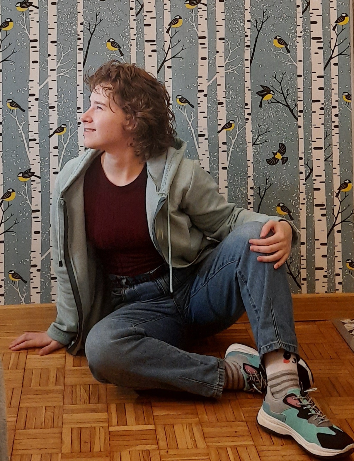 Joanna Puchalska siedzi na drewnianej podłodze. Joanna jest młodą kobietą z kręconymi włosami. Ubrana jest w bluze z kapturem, dżinsy i adidasy. W tle ściana w kolorową tapetą z ptakami.