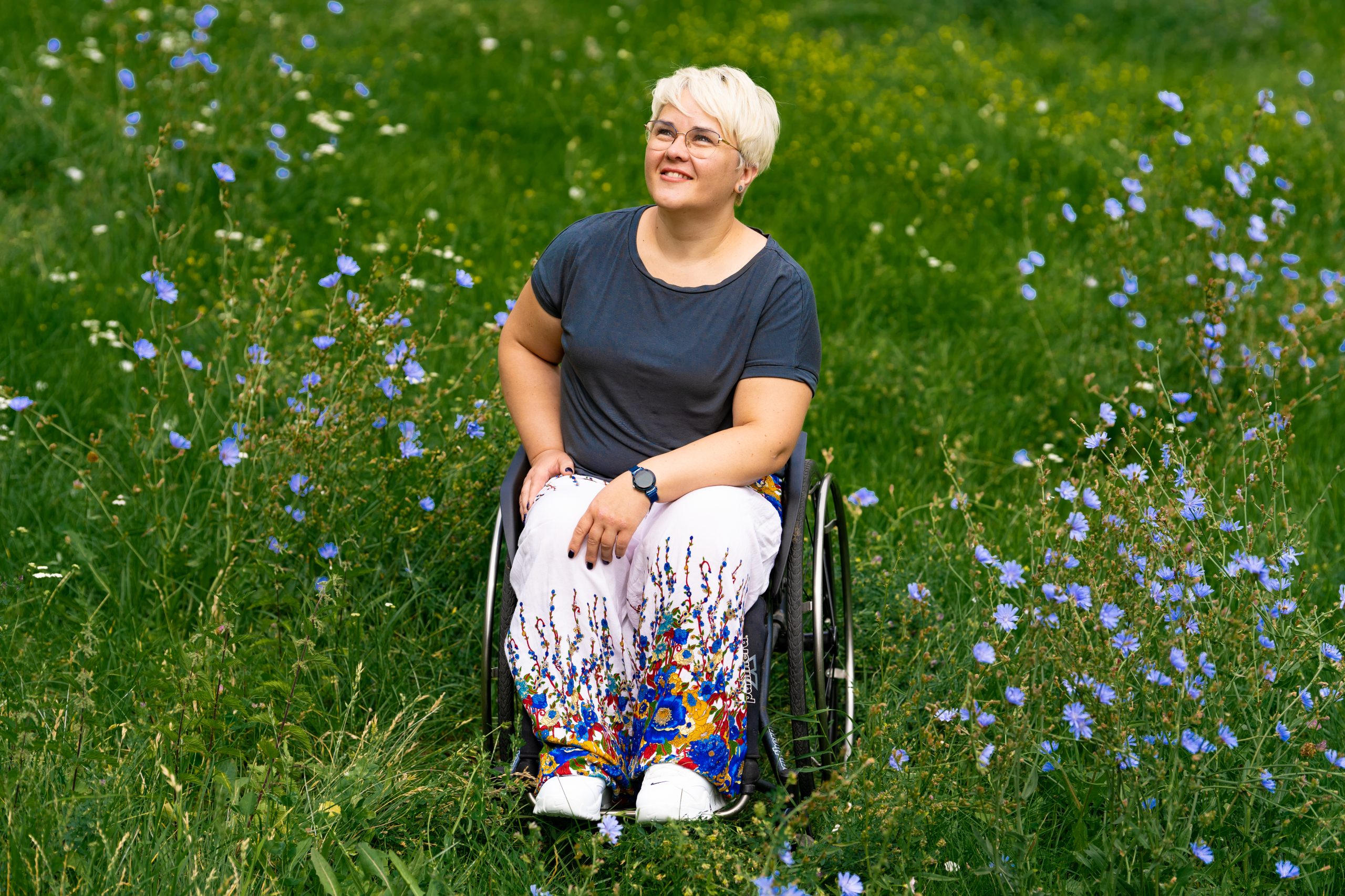 Zdjęcie Justyny Kułakowskiej - około 40 letniej kobiety na wózku. Justyna jest na łace, dookoła rosną polne kwiaty. Uśmiecha się.
