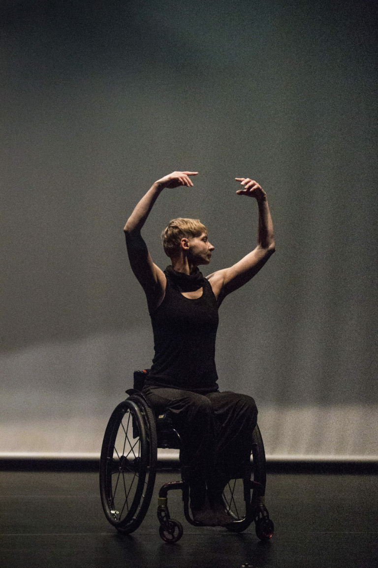 Zdjęcie Tatiany Cholewy - tancerki na wózku, która stoi na scenie. Jest w trakcie spektaklu tańca, ekspresyjnie podnosi ręce.