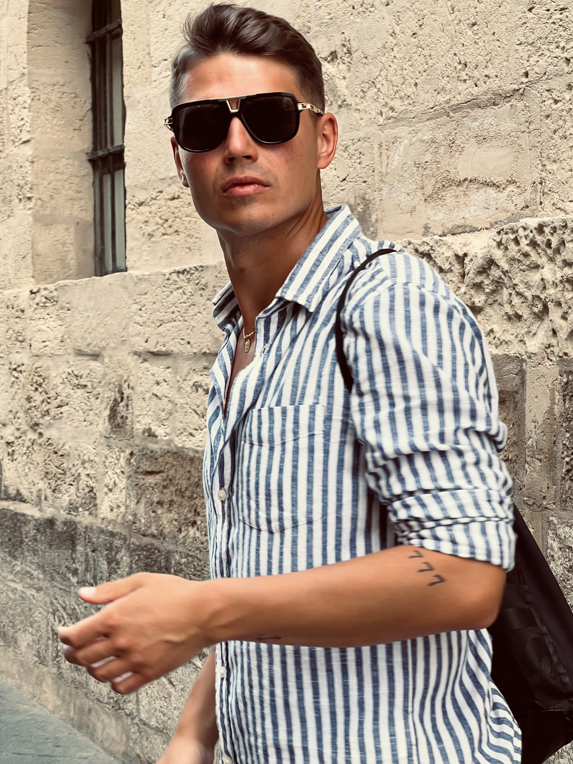 Zdjęcie Flipa, dwudziesto kilku letniego chłopaka. Filip ma na sobie letnią koszulę w paski, plecak i okulary przeciwsłoneczne. Jest opalony. W tle widać ścianę budynku z jasnego piaskowca, który przypomina włoską architekturę.